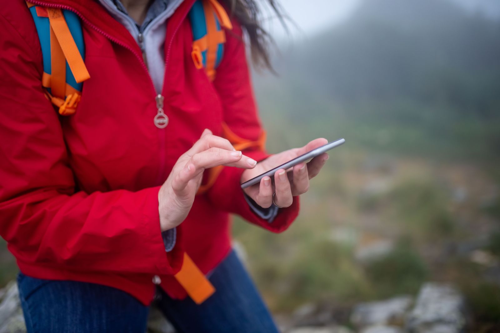 Hands of a hiker in a red jacket checking a smartphone on a misty trail in Portugal