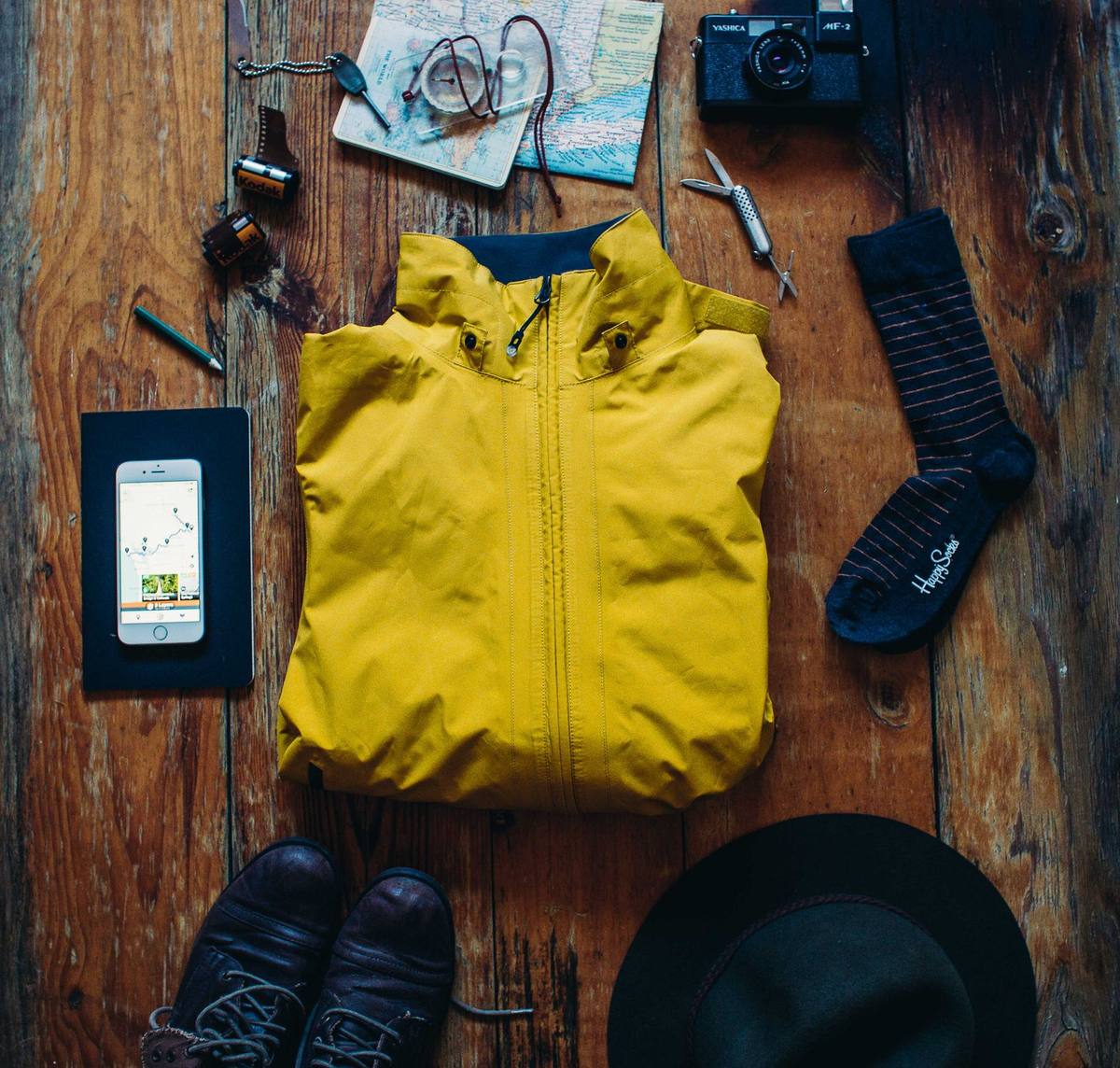 Overhead flat lay of walking gear: leather boots, folded map, camera, jacket, hat and notebook ready for packing